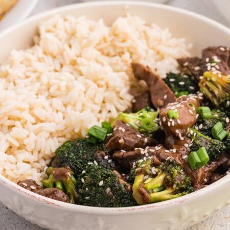Serving bowl with beef and broccoli stir fry and steamed white rice, garnished with sesame seeds and green onions, with spring rolls and side dishes partially visible in the background.