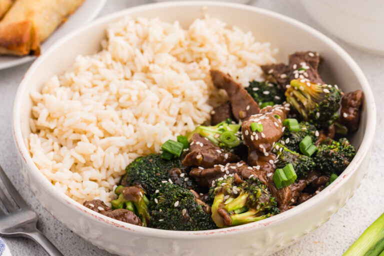 Serving bowl with beef and broccoli stir fry and steamed white rice, garnished with sesame seeds and green onions, with spring rolls and side dishes partially visible in the background.