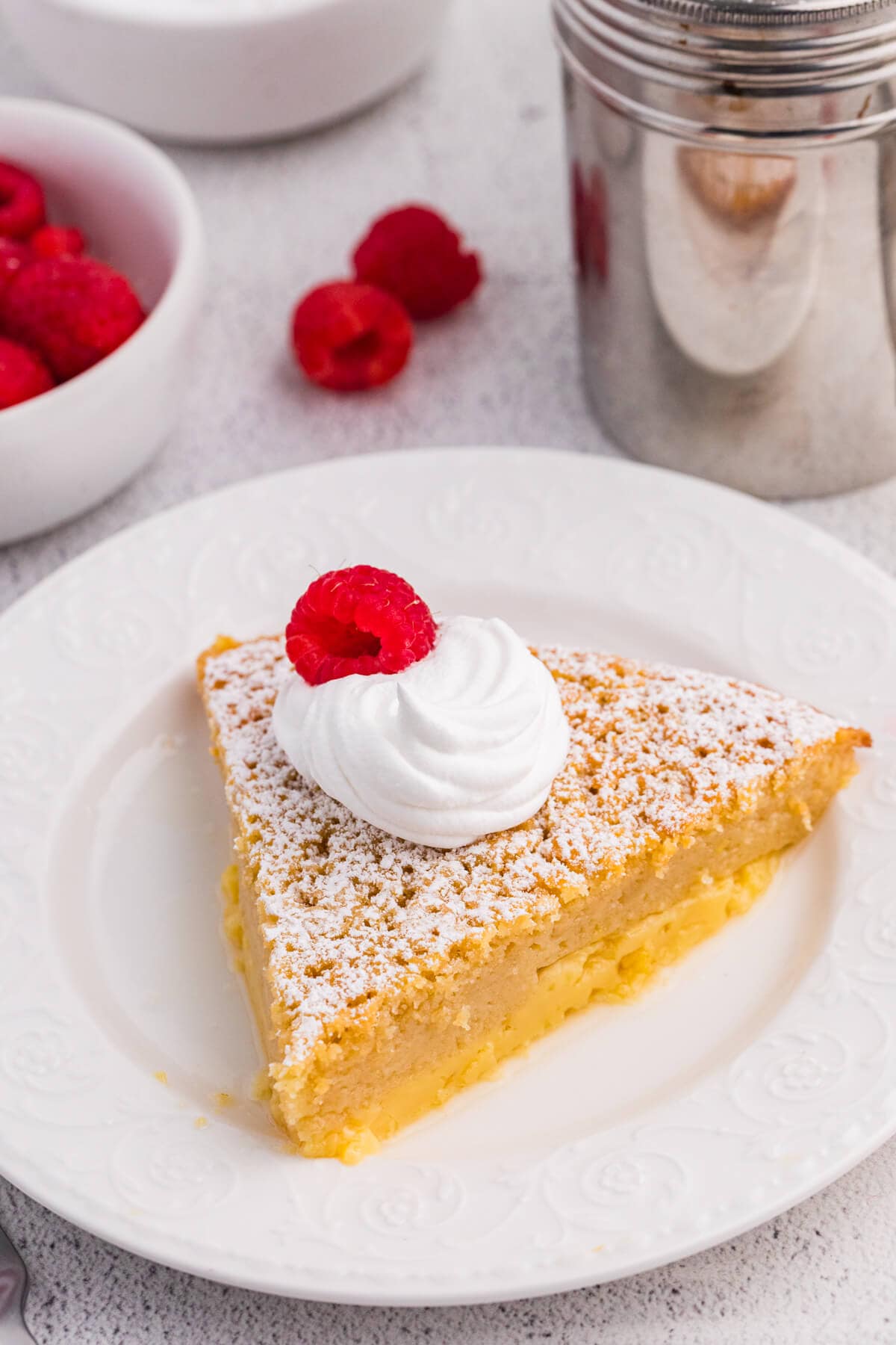 Slice of impossible buttermilk pie topped with whipped cream and a fresh raspberry on a white dessert plate.