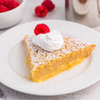 Single slice of impossible buttermilk pie garnished with whipped cream and raspberry, served on a white plate with fresh raspberries nearby.