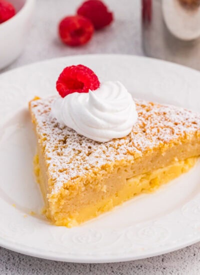 Single slice of impossible buttermilk pie garnished with whipped cream and raspberry, served on a white plate with fresh raspberries nearby.