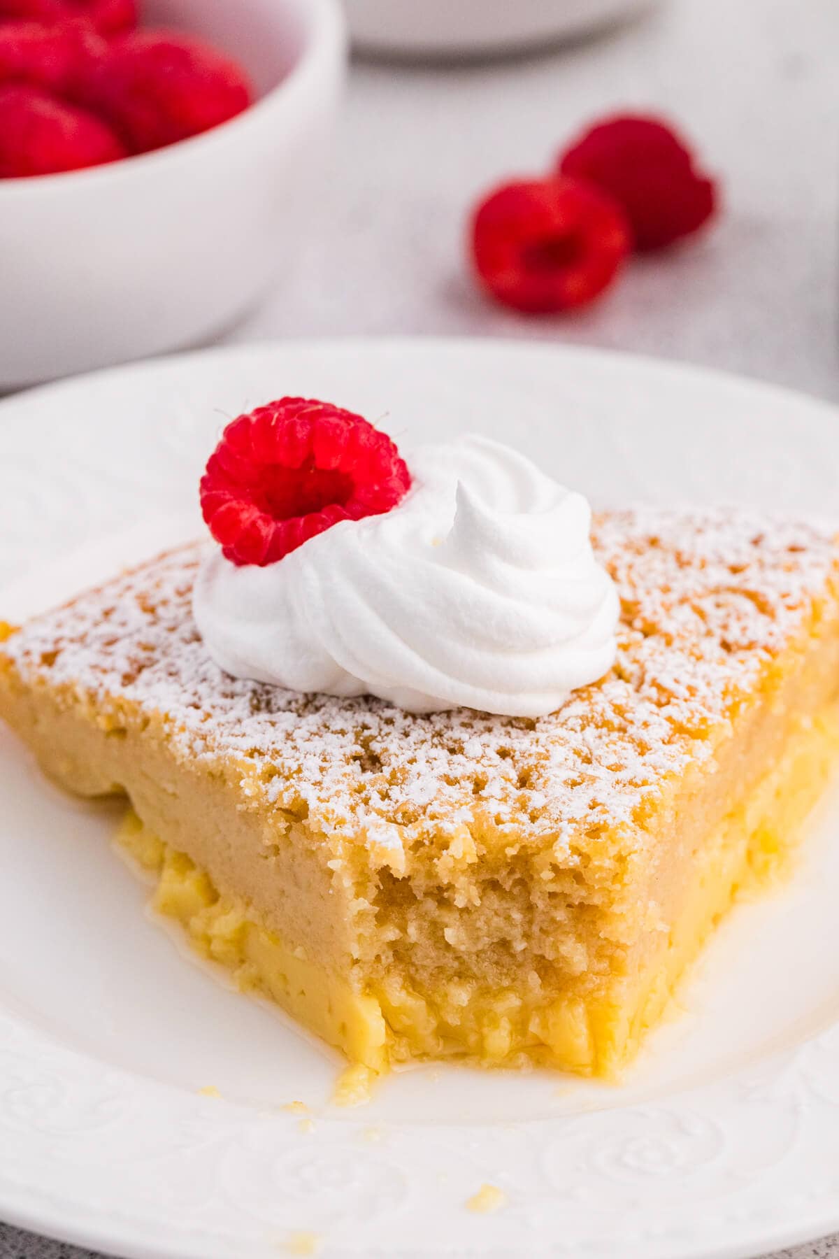 Close-up of impossible buttermilk pie slice with creamy custard layer, topped with whipped cream, powdered sugar, and a raspberry.