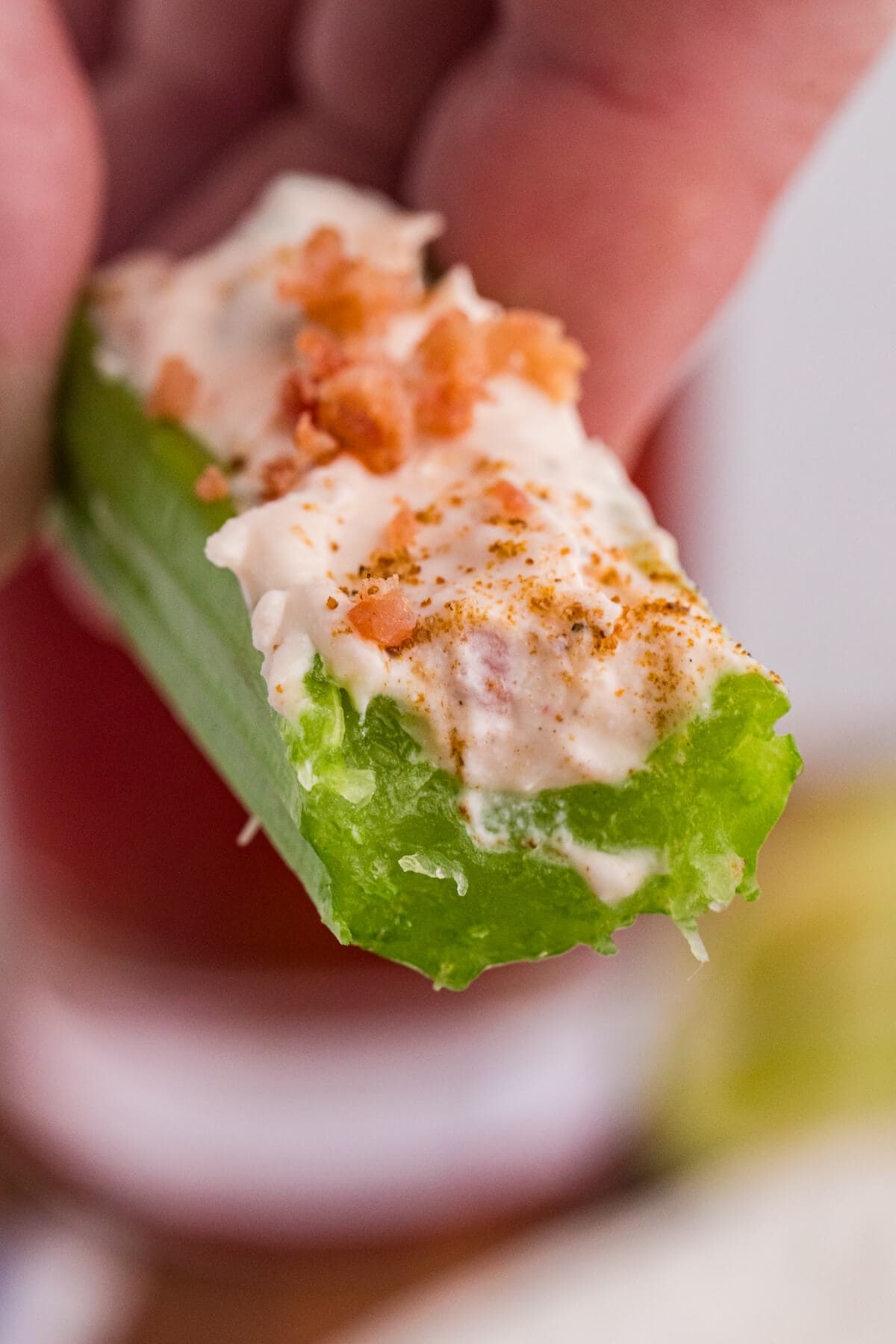 Hand holding a Caesar stuffed celery stick topped with creamy filling, bacon bits, and savory seasoning.