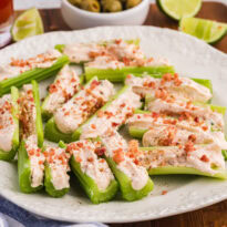 Platter of Caesar stuffed celery appetizers topped with bacon bits, served with olives, lime wedges, and dip.
