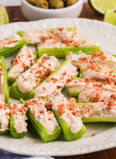 Platter of Caesar stuffed celery appetizers topped with bacon bits, served with olives, lime wedges, and dip.