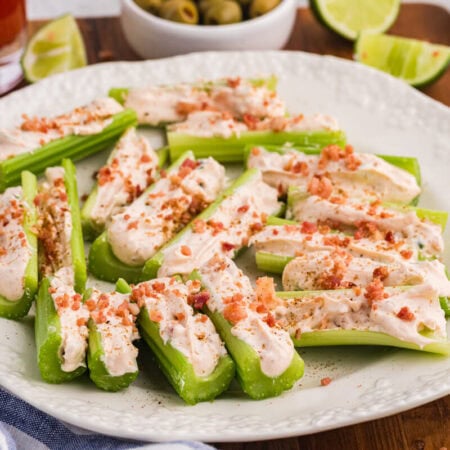 Platter of Caesar stuffed celery appetizers topped with bacon bits, served with olives, lime wedges, and dip.