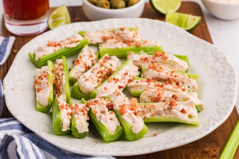 Platter of Caesar stuffed celery appetizers topped with bacon bits, served with olives, lime wedges, and dip.