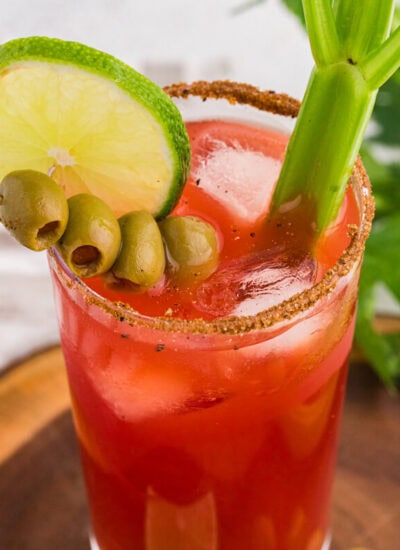 Overhead close-up of a Caesar cocktail with tomato-clam juice, ice, seasoned rim, lime wheel, green olives, and celery garnish, styled on a wooden board with fresh celery and a white cloth background.