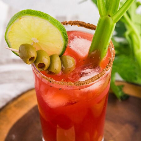Overhead close-up of a Caesar cocktail with tomato-clam juice, ice, seasoned rim, lime wheel, green olives, and celery garnish, styled on a wooden board with fresh celery and a white cloth background.