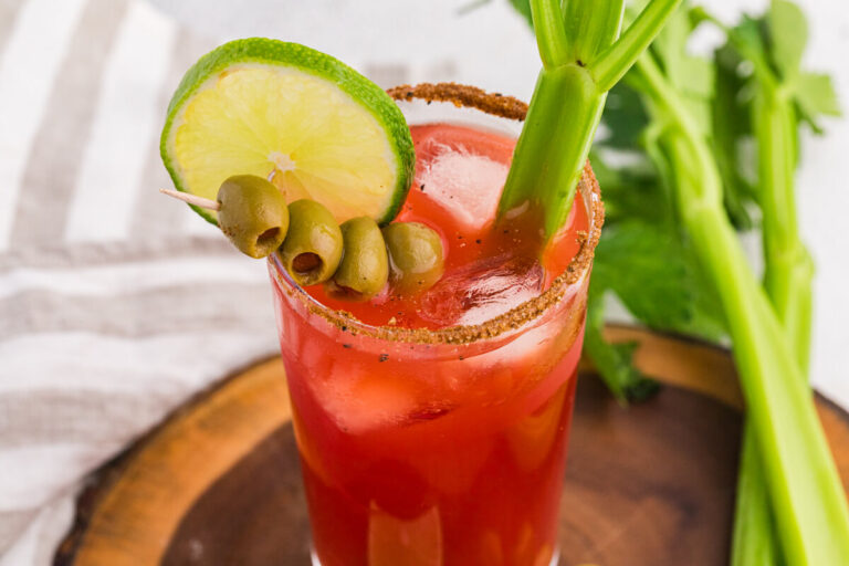 Overhead close-up of a Caesar cocktail with tomato-clam juice, ice, seasoned rim, lime wheel, green olives, and celery garnish, styled on a wooden board with fresh celery and a white cloth background.