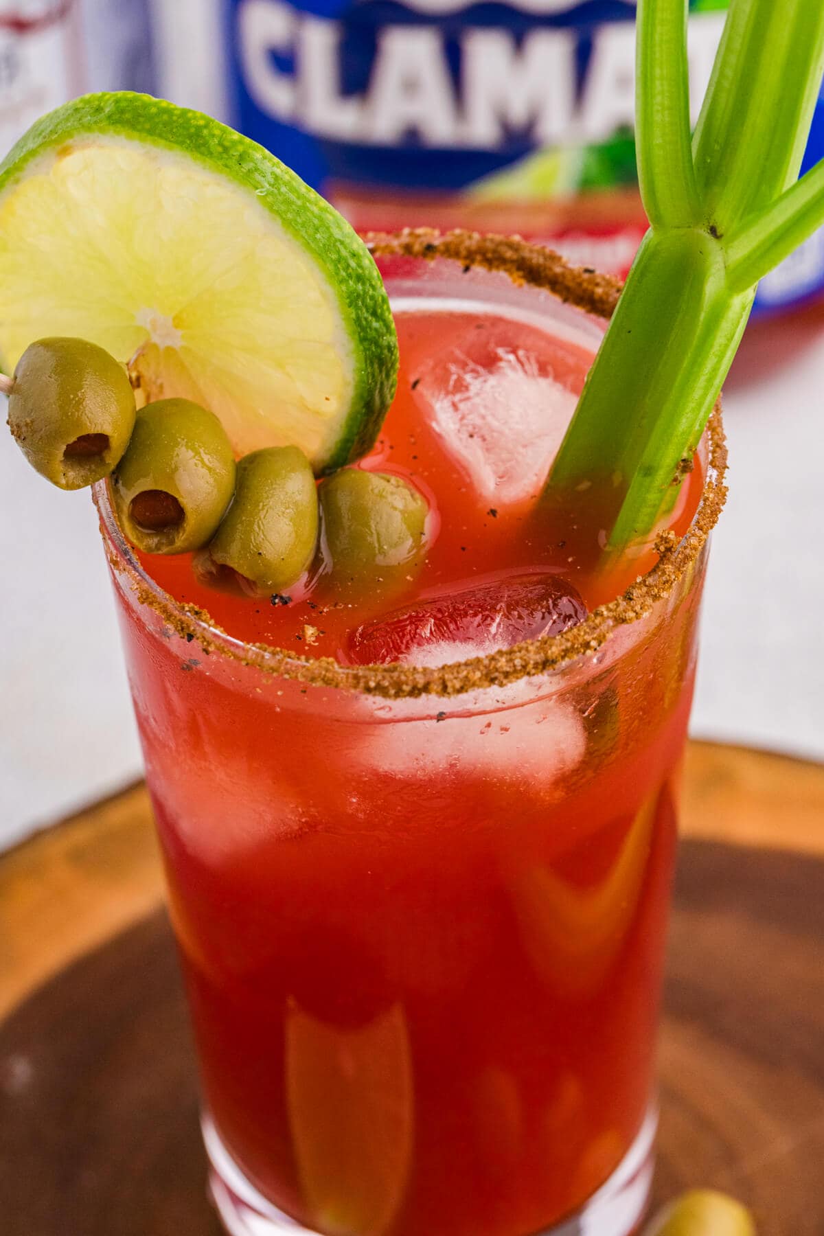 Close-up of a Caesar cocktail with red tomato-clam juice, ice cubes, spiced rim, lime slice, green olives, and celery stalk garnish, with a blurred Clamato bottle behind the glass.