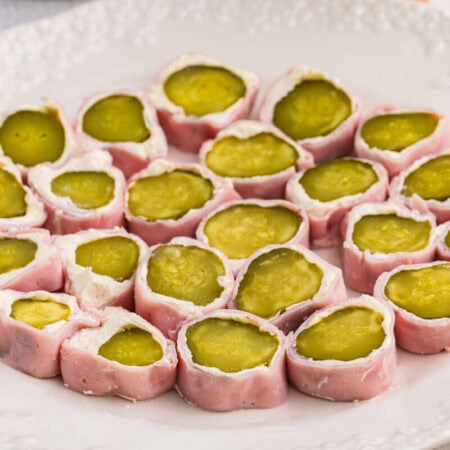 Plate of ham and pickle roll ups sliced into bite-sized appetizers, featuring dill pickles wrapped in cream cheese and deli ham on a white serving plate.