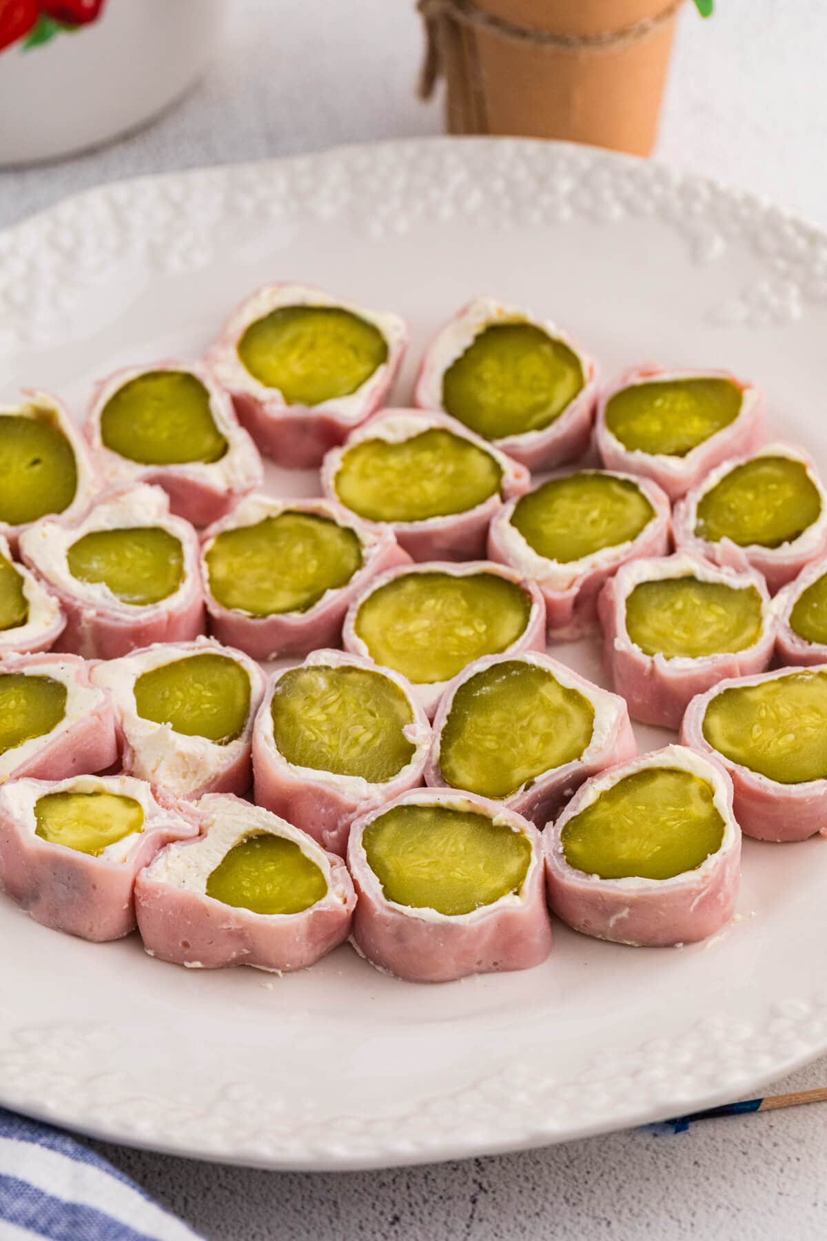 Vertical image of ham and pickle roll ups served on a white textured plate, showing sliced dill pickles wrapped with cream cheese and ham.