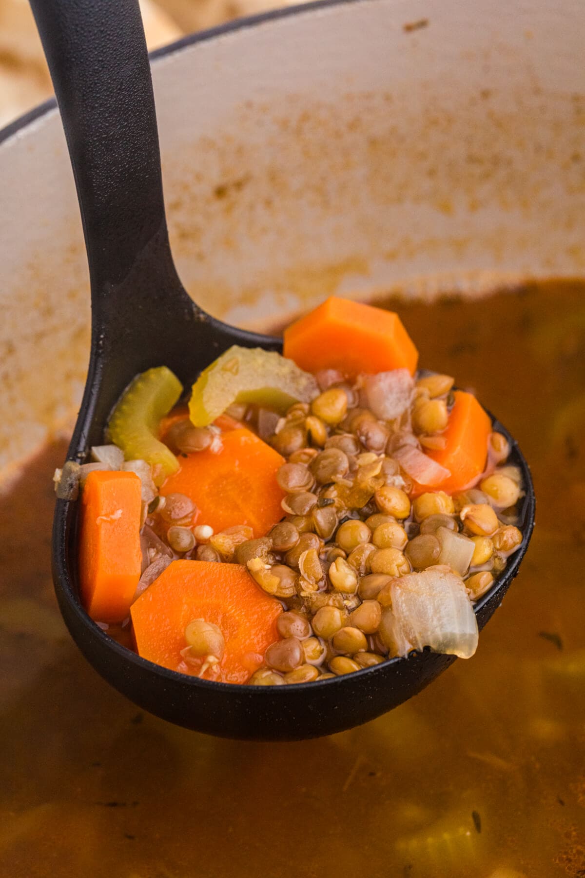 Close-up of a ladle lifting lentil soup with chunks of carrot, celery, and onion in a light brown broth.