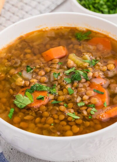 Top view of a bowl of lentil soup with visible vegetables and herbs, served with bread and a spoon on the side.