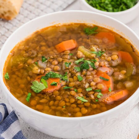 Top view of a bowl of lentil soup with visible vegetables and herbs, served with bread and a spoon on the side.