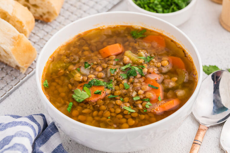 Top view of a bowl of lentil soup with visible vegetables and herbs, served with bread and a spoon on the side.