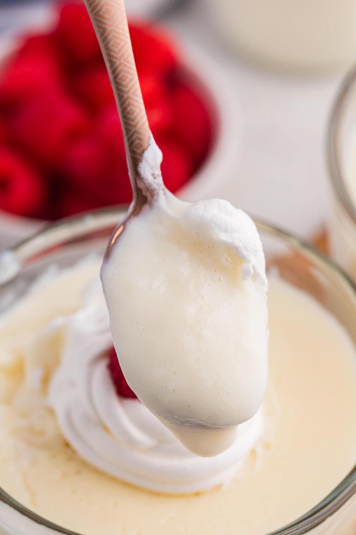 Spoon lifting a soft, airy scoop of vanilla milk pudding from a glass dessert, showing its light, mousse-like texture with raspberries blurred in the background.