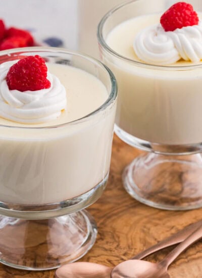 Two glass dessert cups filled with smooth vanilla milk pudding, each topped with a swirl of whipped cream and a fresh raspberry, set on a wooden board with a small bottle of milk and a bowl of raspberries in the background.