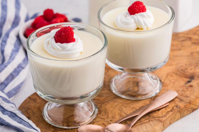 Two glass dessert cups filled with smooth vanilla milk pudding, each topped with a swirl of whipped cream and a fresh raspberry, set on a wooden board with a small bottle of milk and a bowl of raspberries in the background.