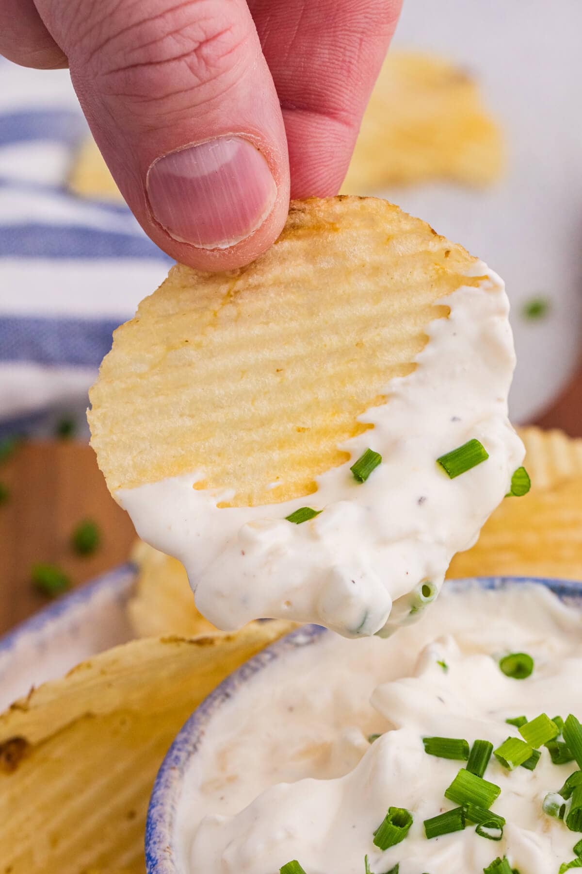 Hand holding ridged chip dipped in thick onion dip with visible chives.