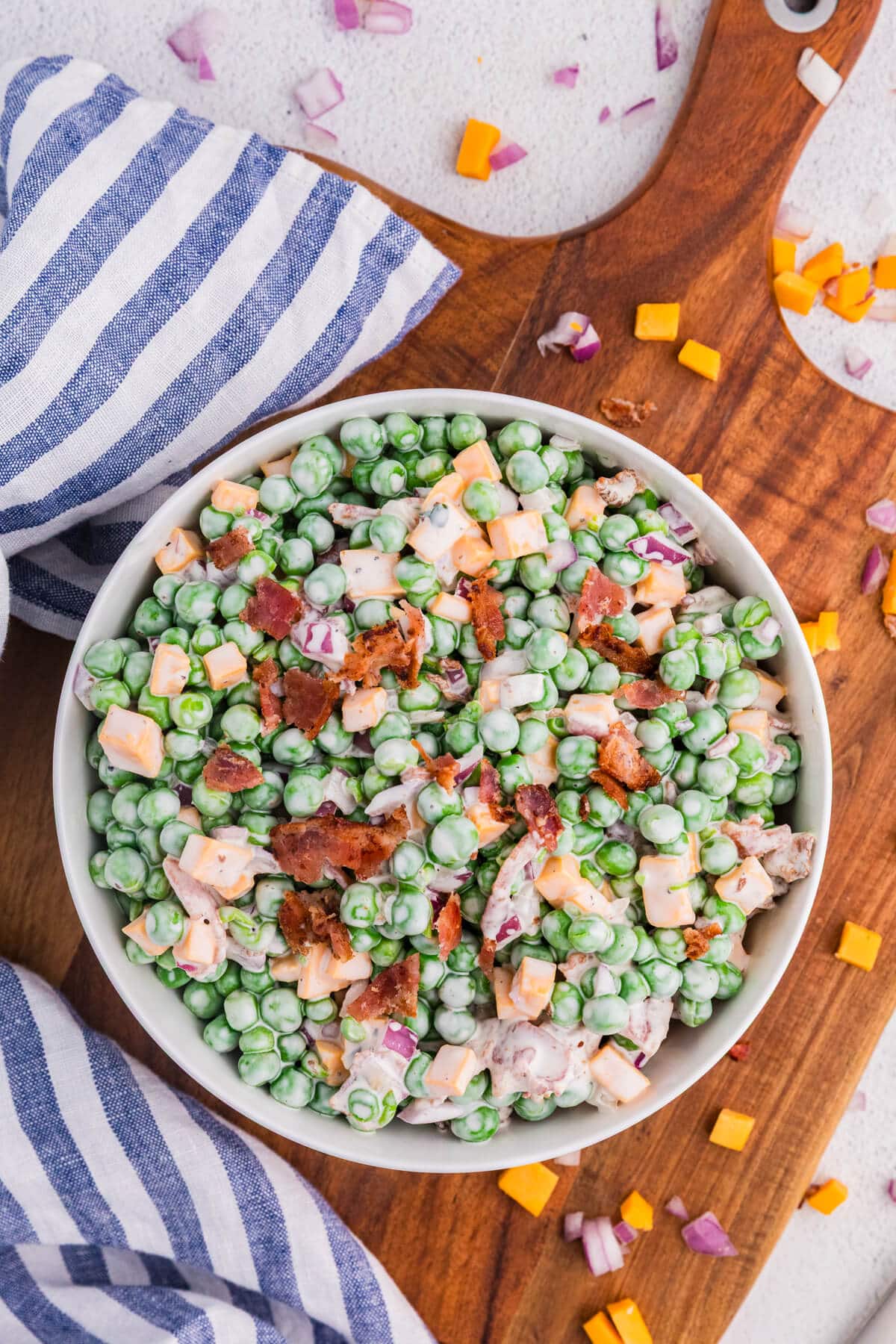 Overhead view of creamy pea salad with green peas, cheddar cheese cubes, crispy bacon, and red onion in a white bowl on a wooden cutting board with striped cloth, fresh summer side dish.