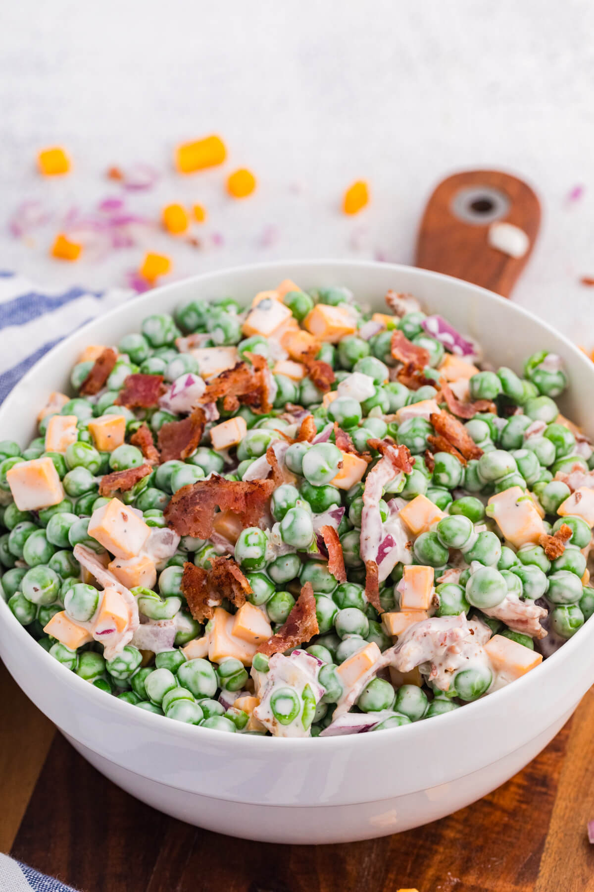 Close-up angled shot of pea salad with creamy dressing, green peas, cheddar cubes, bacon bits, and diced red onions in a white bowl, rustic kitchen background with scattered ingredients.