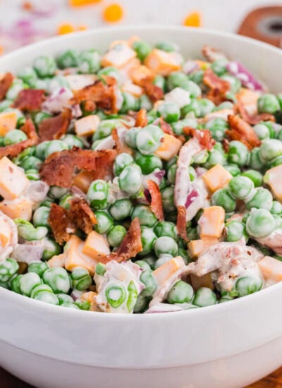 Side view of creamy pea salad in a white bowl featuring green peas, cheese cubes, bacon, and red onion, served on a wooden board with soft blurred background.