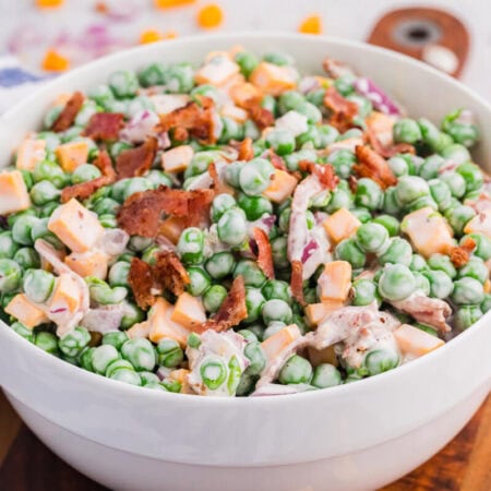 Side view of creamy pea salad in a white bowl featuring green peas, cheese cubes, bacon, and red onion, served on a wooden board with soft blurred background.