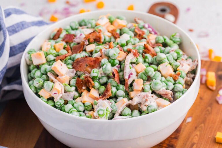 Side view of creamy pea salad in a white bowl featuring green peas, cheese cubes, bacon, and red onion, served on a wooden board with soft blurred background.