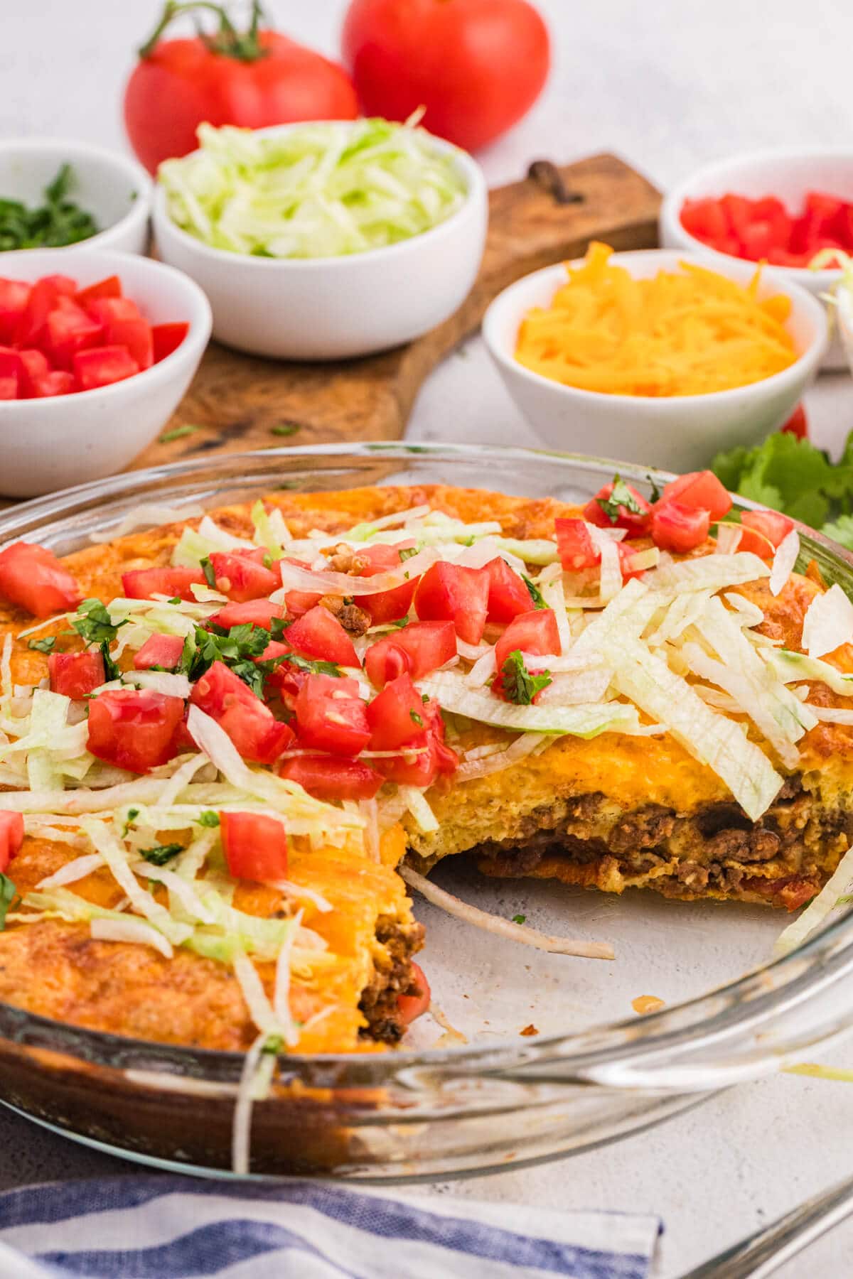 Taco pie with a slice removed, showing layers of seasoned ground beef and fluffy crust, topped with lettuce, tomatoes, and cilantro.
