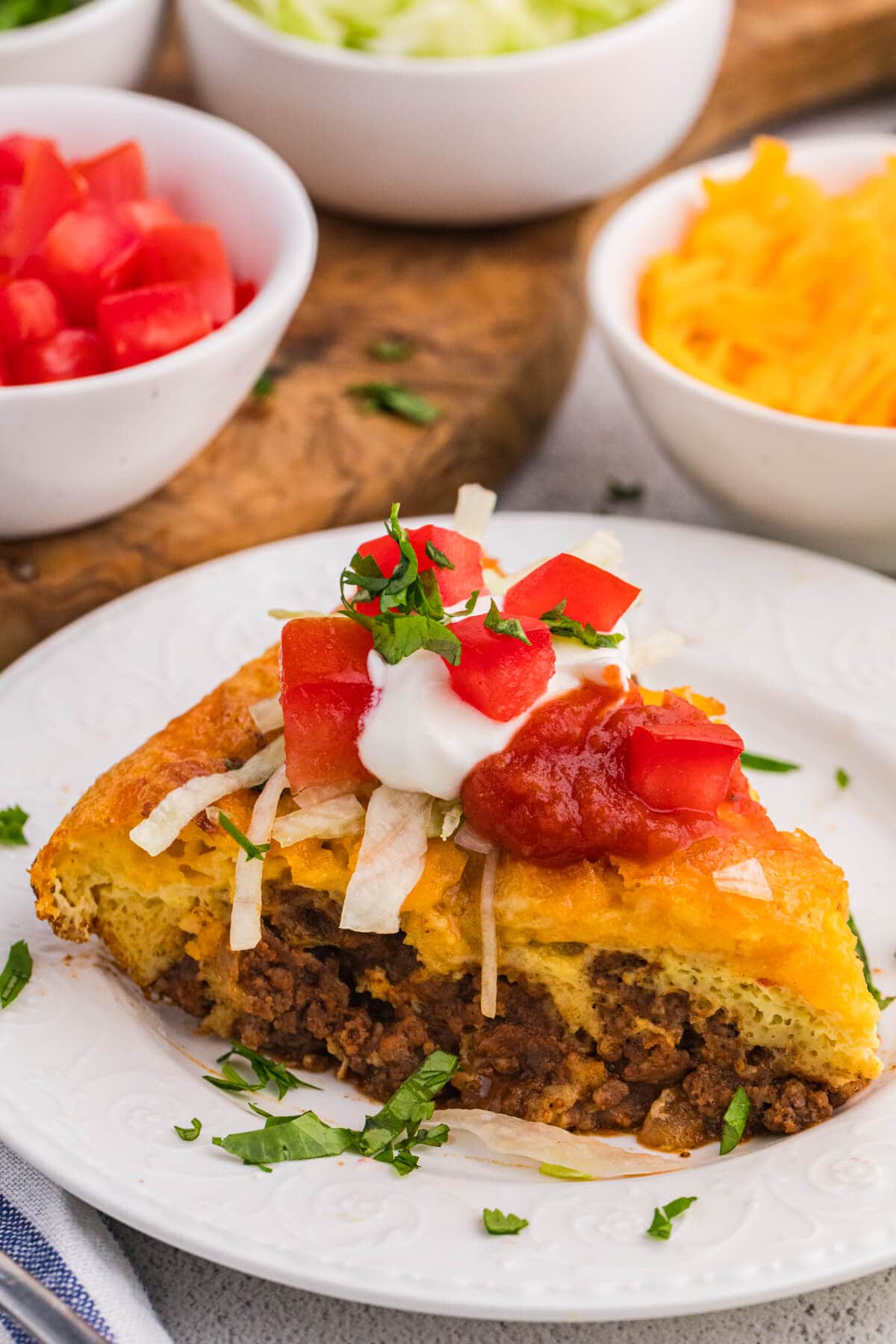 Close-up of a plated taco pie slice with ground beef filling, topped with sour cream, salsa, lettuce, and tomatoes, garnished with chopped herbs.