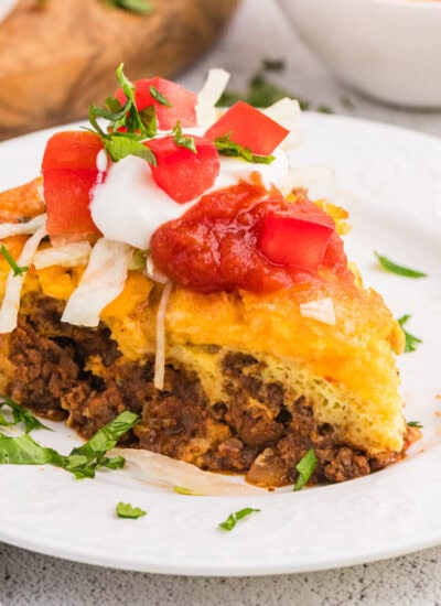Single slice of taco pie on a white plate topped with sour cream, salsa, diced tomatoes, and lettuce, with fresh ingredients in the background.