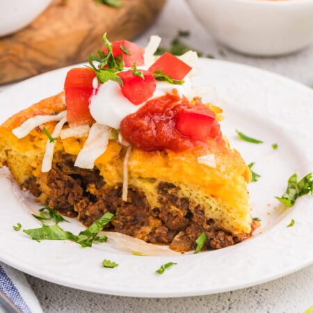 Single slice of taco pie on a white plate topped with sour cream, salsa, diced tomatoes, and lettuce, with fresh ingredients in the background.