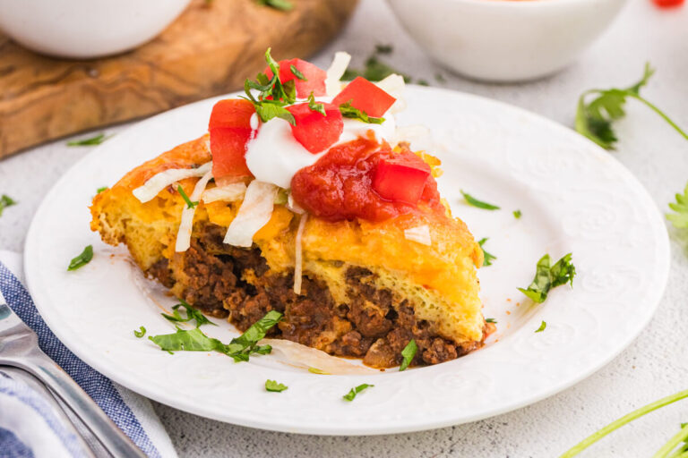 Single slice of taco pie on a white plate topped with sour cream, salsa, diced tomatoes, and lettuce, with fresh ingredients in the background.