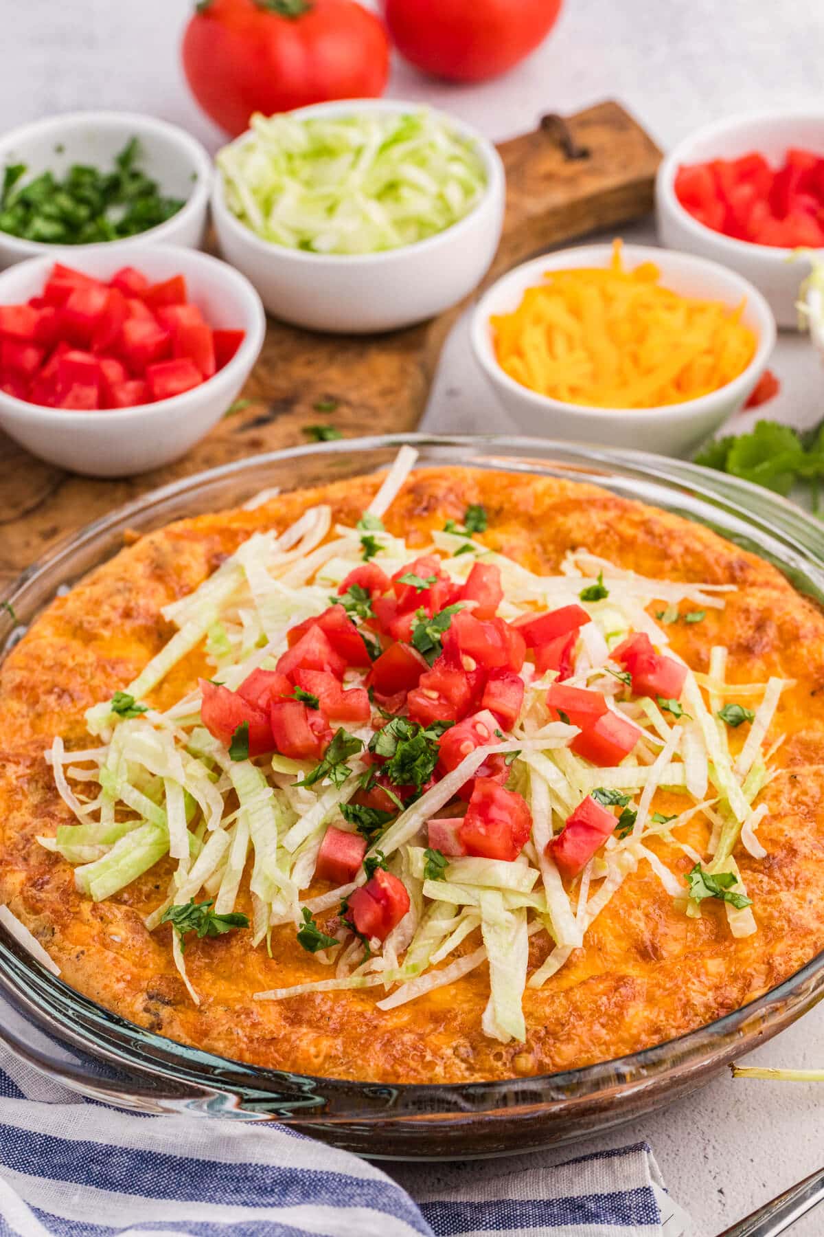 Angled close-up of a golden taco pie topped with lettuce, tomatoes, and herbs, with fresh ingredients in small bowls blurred in the background.
