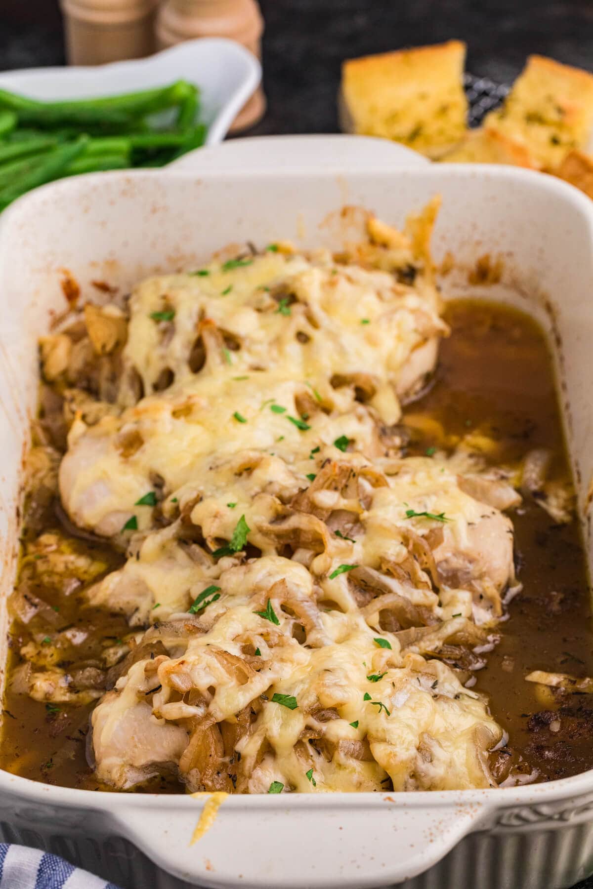 Close-up of French onion chicken bake in a white baking dish with melted cheese, caramelized onions, savory sauce, green beans, and garlic bread in the background.