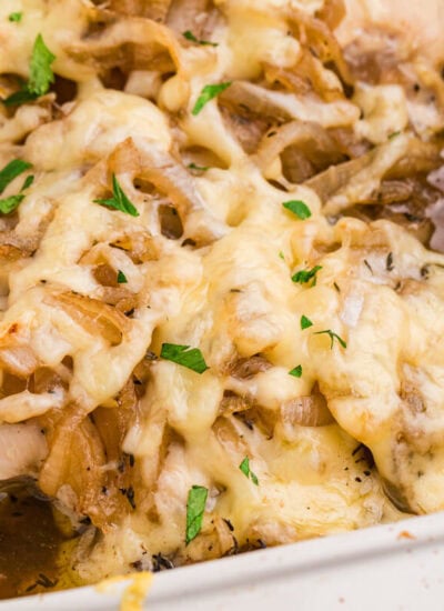 French onion chicken bake topped with melted cheese, caramelized onions, parsley, and herbs in a rich brown pan sauce inside a white casserole dish.