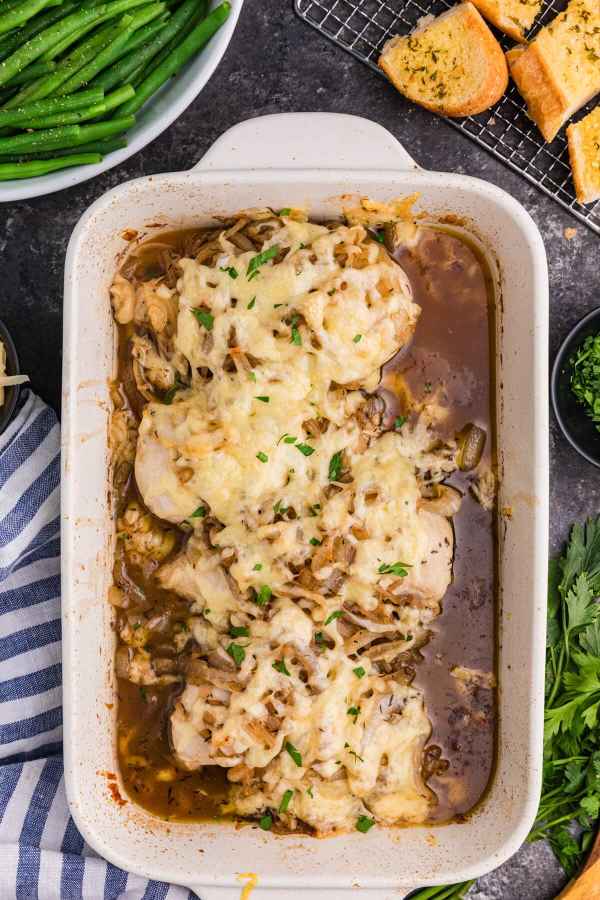 French onion chicken bake in a white casserole dish, topped with melted cheese, caramelized onions, herbs, and pan sauce, served with green beans and garlic bread.