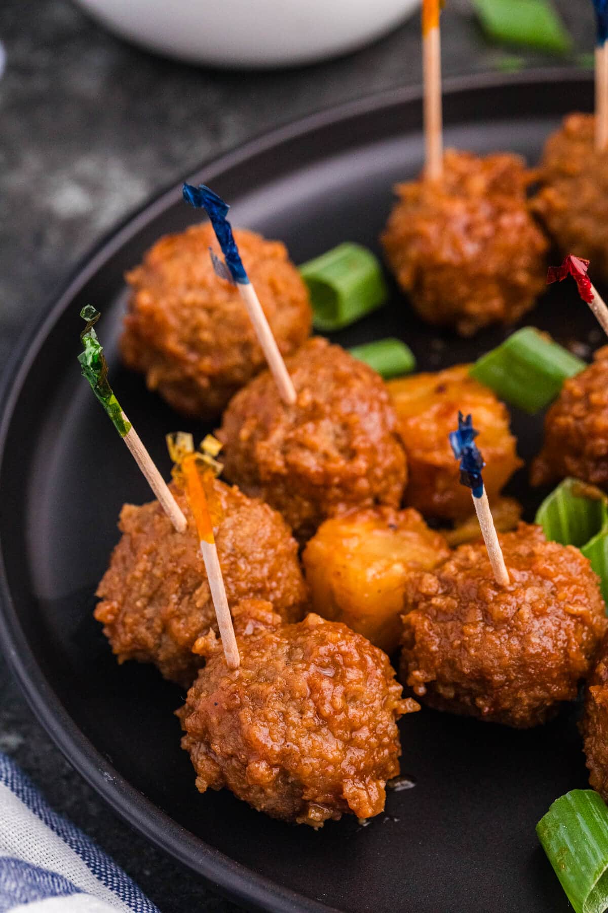 Pineapple BBQ meatballs served as appetizers on a black plate with toothpicks, pineapple chunks, and sliced green onions.