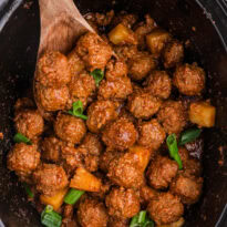 Pineapple BBQ meatballs in a black slow cooker, coated in tangy barbecue sauce and topped with pineapple chunks and green onions.