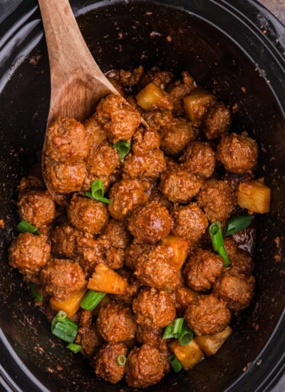 Pineapple BBQ meatballs in a black slow cooker, coated in tangy barbecue sauce and topped with pineapple chunks and green onions.