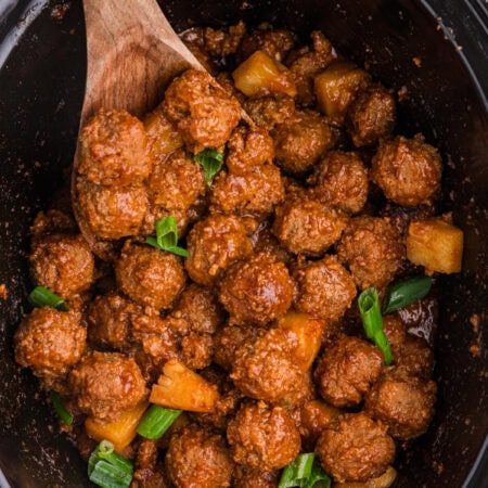 Pineapple BBQ meatballs in a black slow cooker, coated in tangy barbecue sauce and topped with pineapple chunks and green onions.