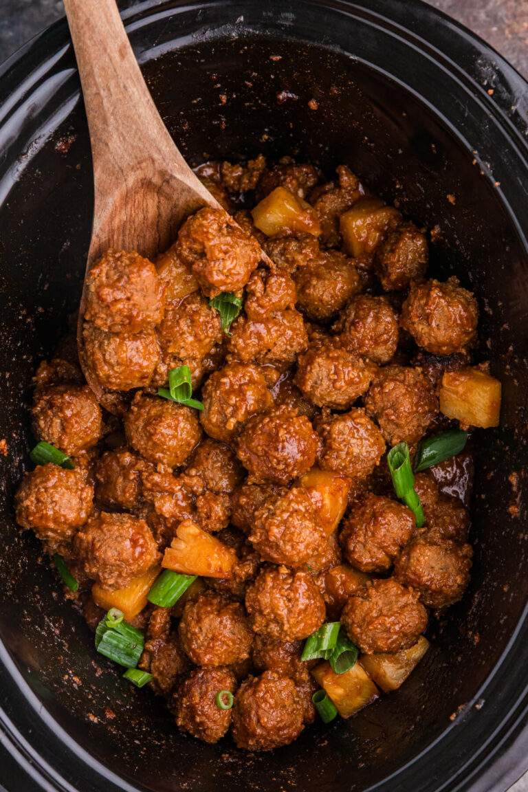 Pineapple BBQ meatballs in a black slow cooker, coated in tangy barbecue sauce and topped with pineapple chunks and green onions.