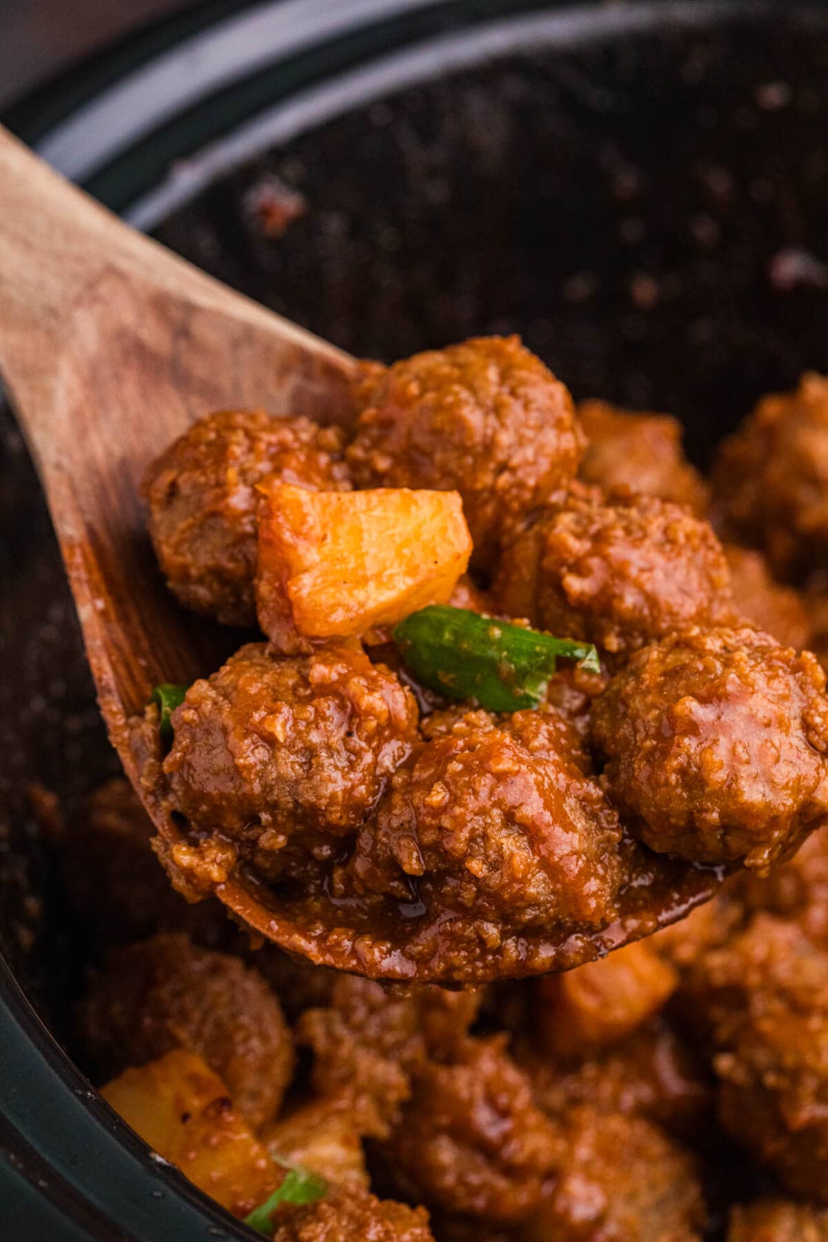 Close-up of pineapple BBQ meatballs on a wooden spoon, glazed with barbecue sauce and garnished with pineapple and green onion.