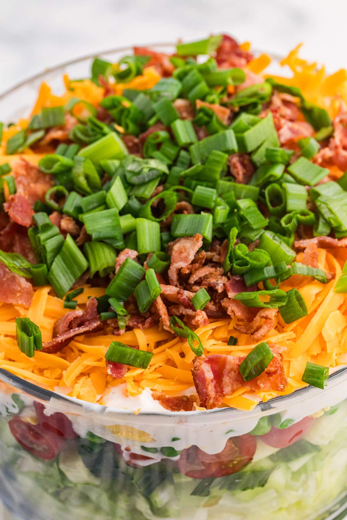 Close-up of seven layer salad topped with shredded cheddar cheese, crispy bacon, and fresh green onions in a glass serving bowl.