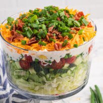 Seven layer salad in a clear trifle bowl with lettuce, cucumber, tomatoes, peas, creamy dressing, cheddar cheese, bacon, and green onions.