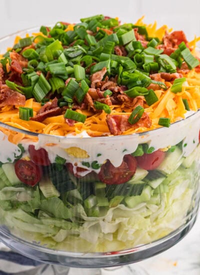 Seven layer salad in a clear trifle bowl with lettuce, cucumber, tomatoes, peas, creamy dressing, cheddar cheese, bacon, and green onions.