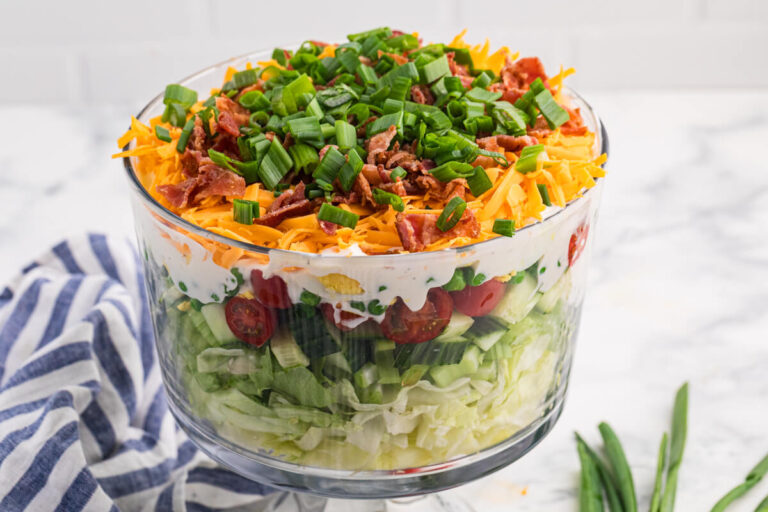 Seven layer salad in a clear trifle bowl with lettuce, cucumber, tomatoes, peas, creamy dressing, cheddar cheese, bacon, and green onions.