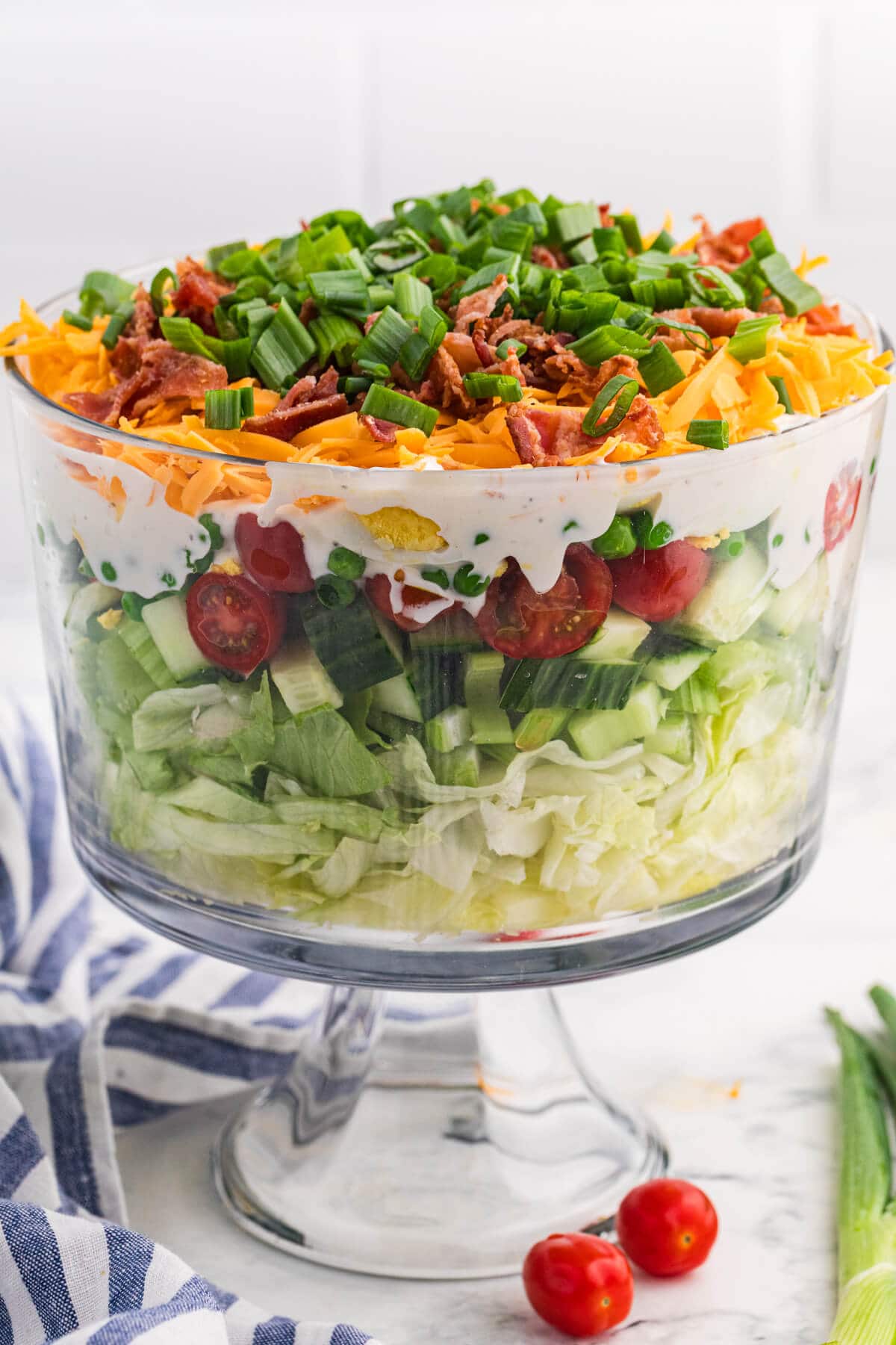 Seven layer salad served in a pedestal glass bowl with visible vegetable layers, creamy dressing, cheddar cheese, bacon, and green onion topping.